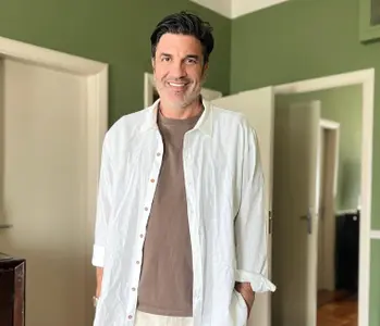 O apresentador e chef de cozinha Edu Guedes, usando camiseta marrom e camisa branca, sorri, em pé, diante da câmera
