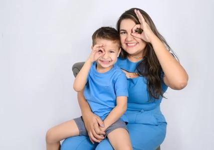 Menino sentado no colo da mãe. Ambos destacam os olhos, com um gesto feito com as mãos, em campanha de conscientização sobre o retinoblastoma, câncer pediátrico